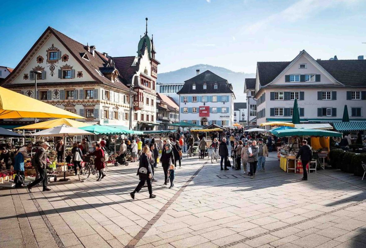 A market with a umber of people. There are stalls in the centre and it is rimmed by quaint buildings.