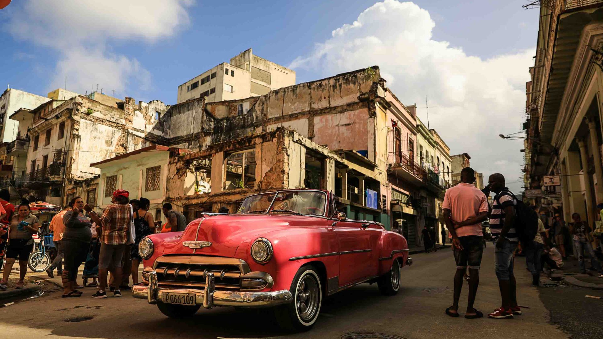 People stand around in the street talking. There's a pink car in the centre of them.