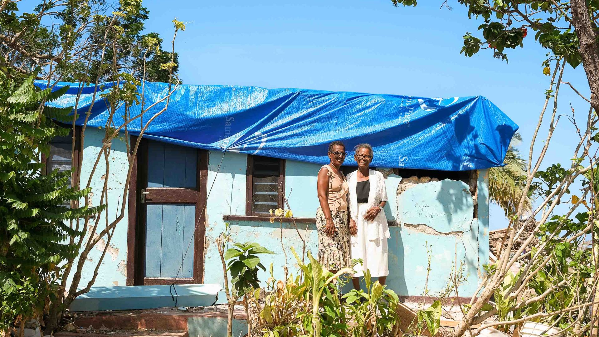 Two women stand in front of a house with a tarpaulin on its roof.