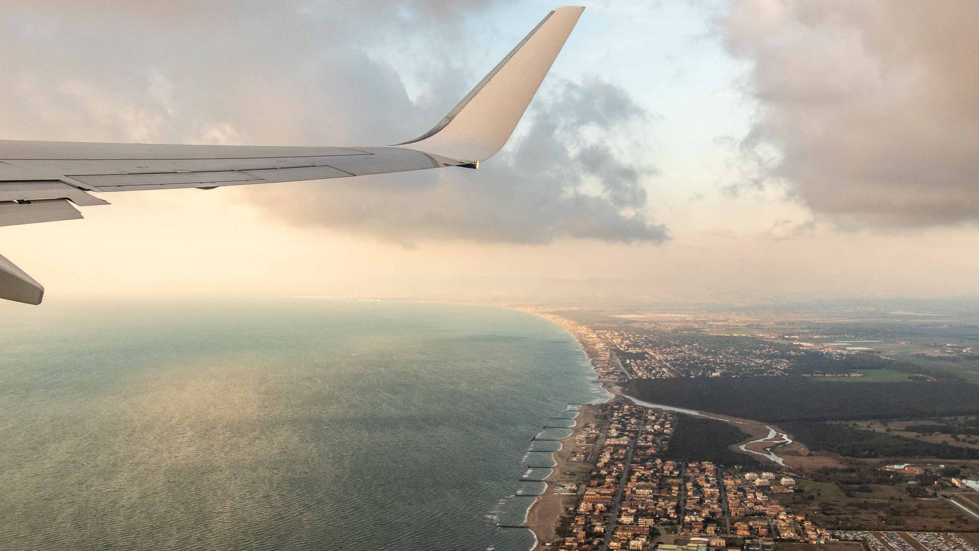 The wing of a plane is seen flying over water and a city.