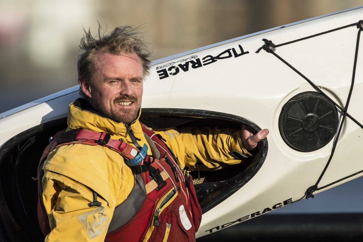 A man wearing a yellow jacket smiles to camera as he holds a kayak.