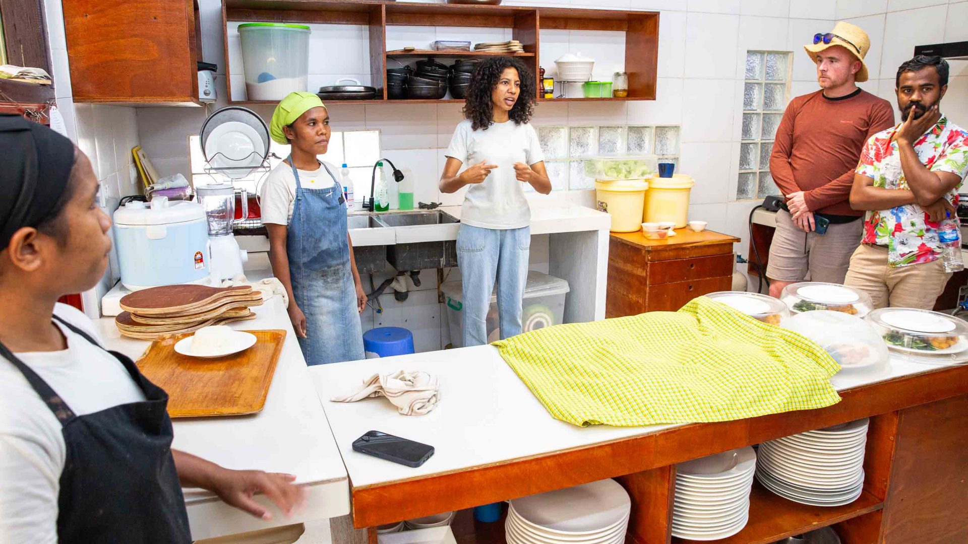 A woman stands in the kitchen of a restaurant where she talks to guests who are standing around.