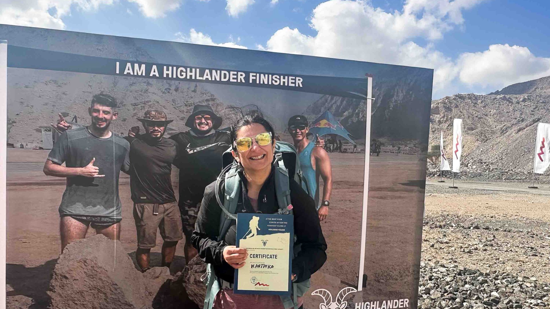 A woman holds a certificate of completion in front of a sign for the Highlander trail.